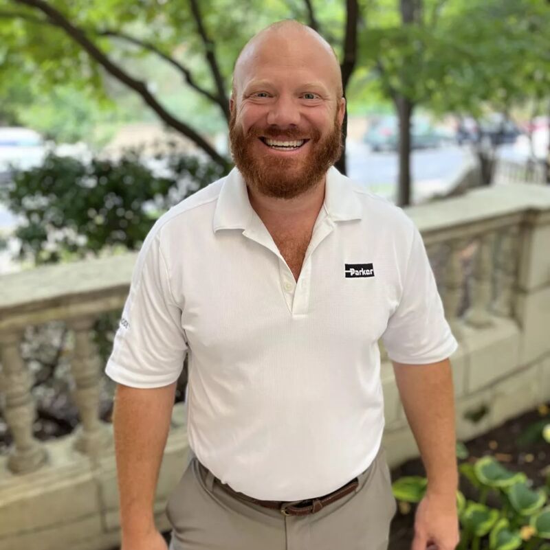The image shows a smiling man with a red beard and a shaved head. He is wearing a white polo shirt with a logo and khaki pants. He is standing outside near a stone wall and greenery. The man appears to be happy and relaxed.